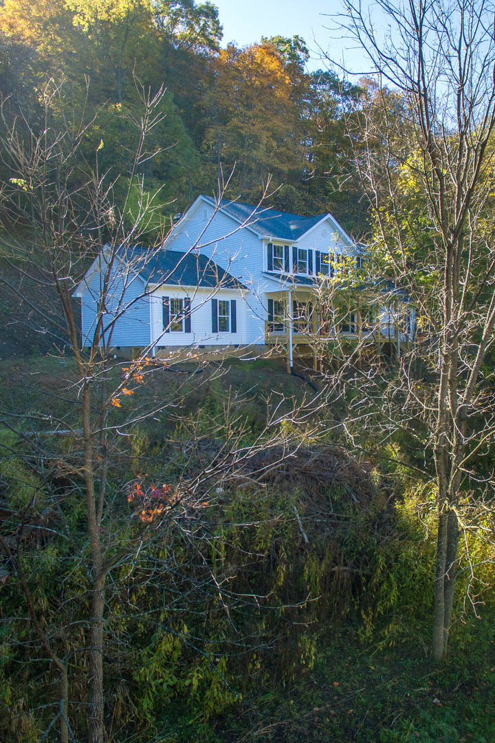 Two-story farmhouse with blue roof, surrounded by leafy and bare trees, grassy lawn, and multiple windows