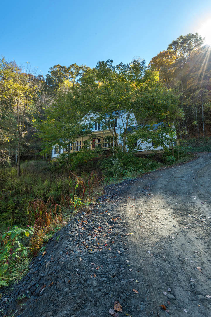 Two-story house with light siding sits atop a grassy hill, surrounded by trees with yellow autumn leaves, dirt road leading up, blue sky overhead