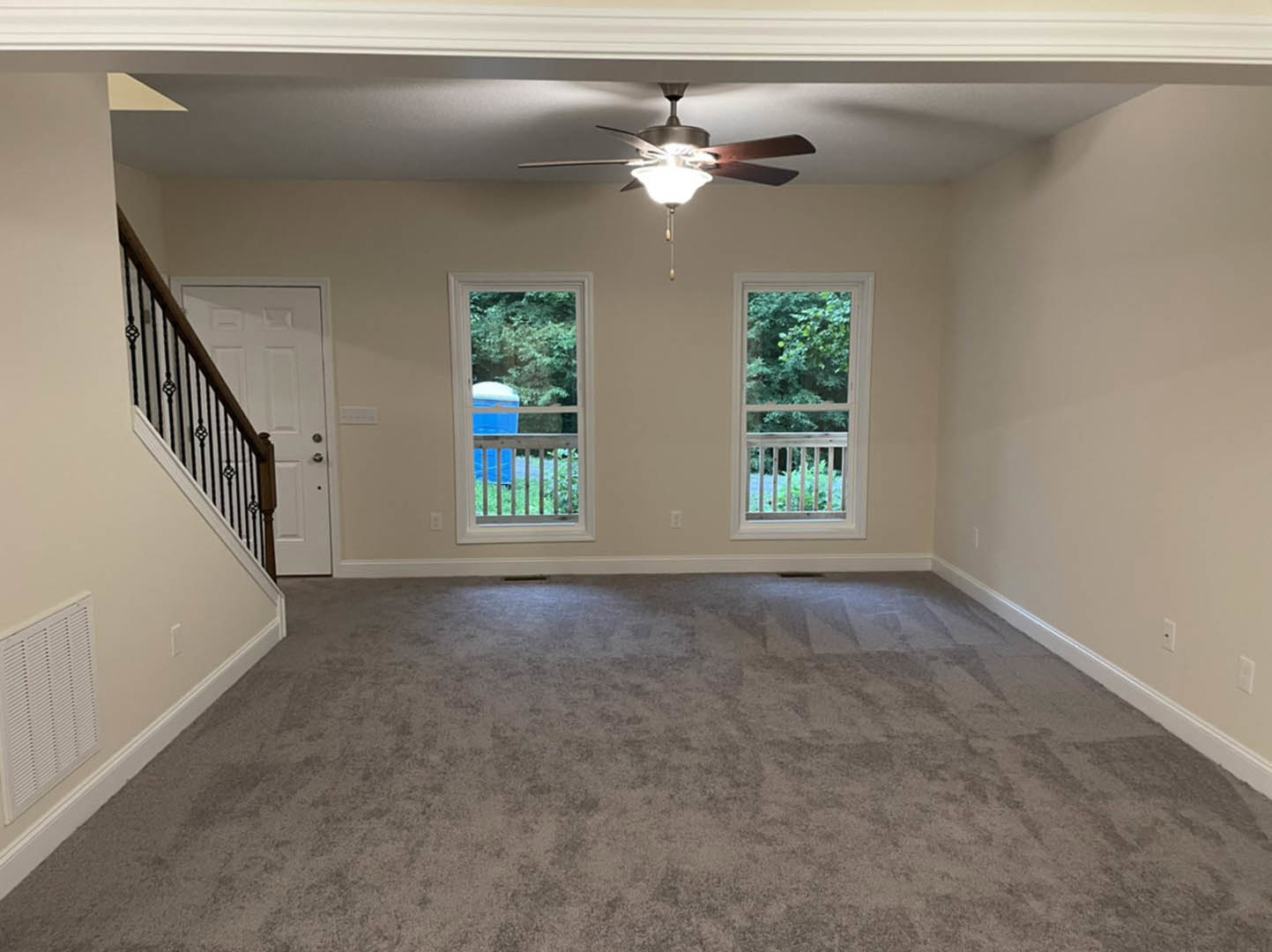 Carpeted living room featuring a ceiling fan with light fixture, two windows with white trim, wall-mounted light, and staircase with wooden railing