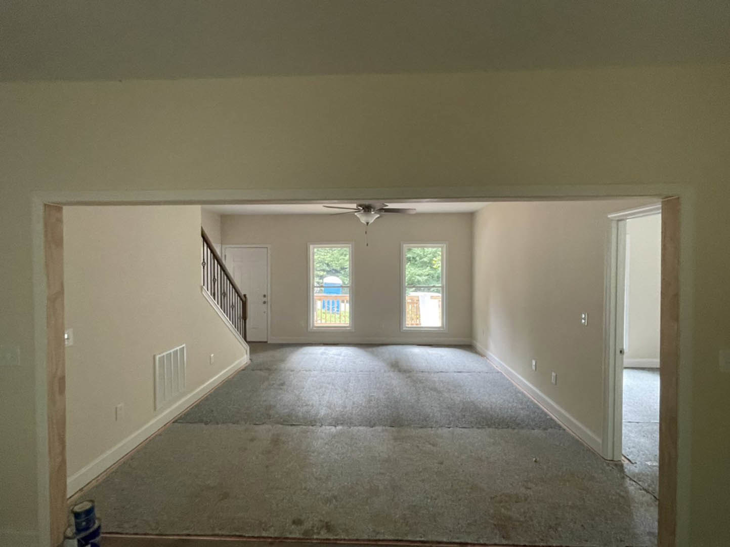 Carpeted living area with white walls, wooden staircase, ceiling fan, large windows overlooking trees, and a blue planter on the balcony