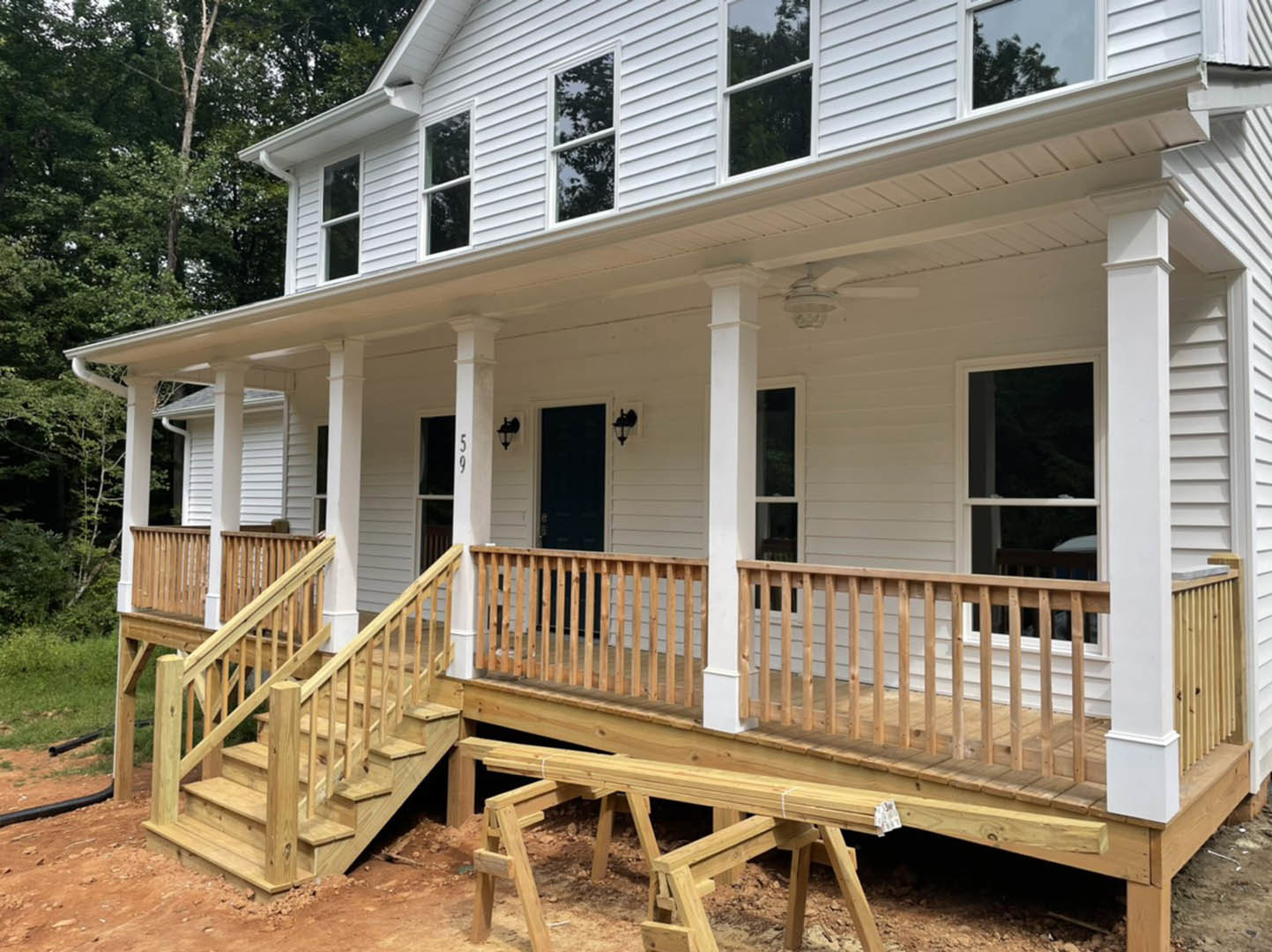 White siding house with covered porch, wooden stairs, white framed windows reflecting trees, white shutters, and wooden deck with bench