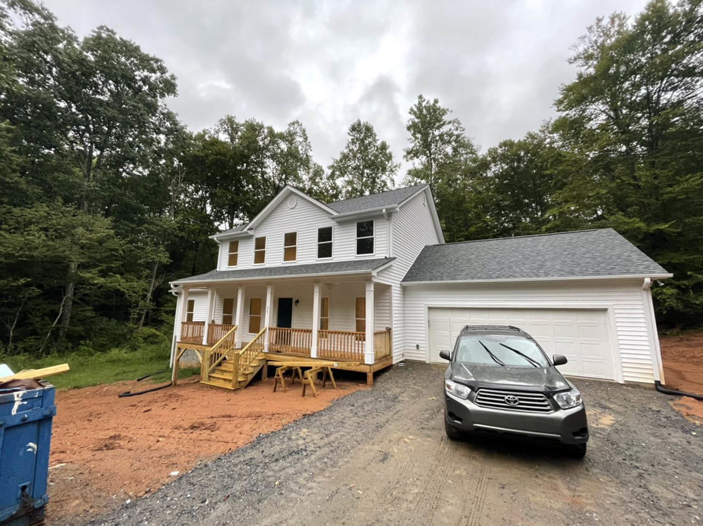 Gray sedan parked on concrete driveway in front of two-story house with blue door, white trim, wooden porch, and deck; leafy trees and cloudy sky in background.