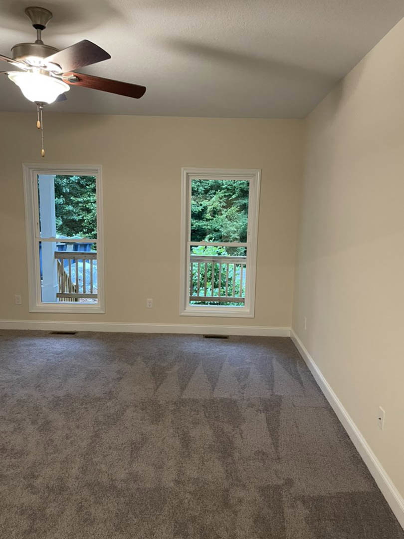 Carpeted bedroom with two large windows overlooking balcony and trees, white plaster walls, ceiling fan with light fixture, floor vent near window railing