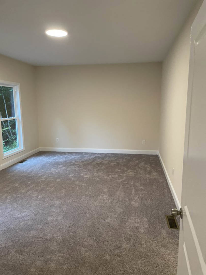 Carpeted room with white walls, white trim, ceiling light fixture, door, window, and wall vent