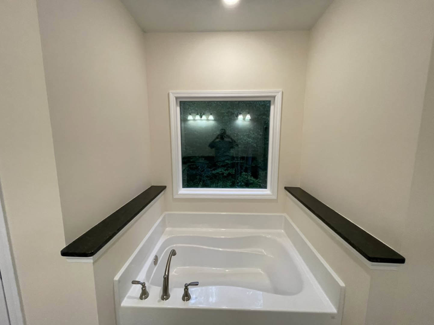 Freestanding white bathtub beneath large window, chrome faucet, black floating shelf on white tile wall, bathroom accessories visible