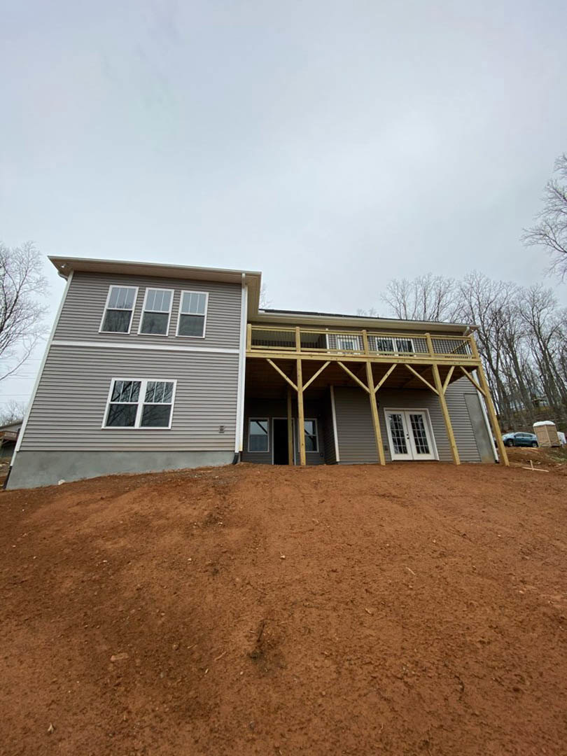 Two-story house with white siding, covered front porch, balcony above, double doors, multiple windows with white frames, set on a dirt hill with trees and cloudy sky in the