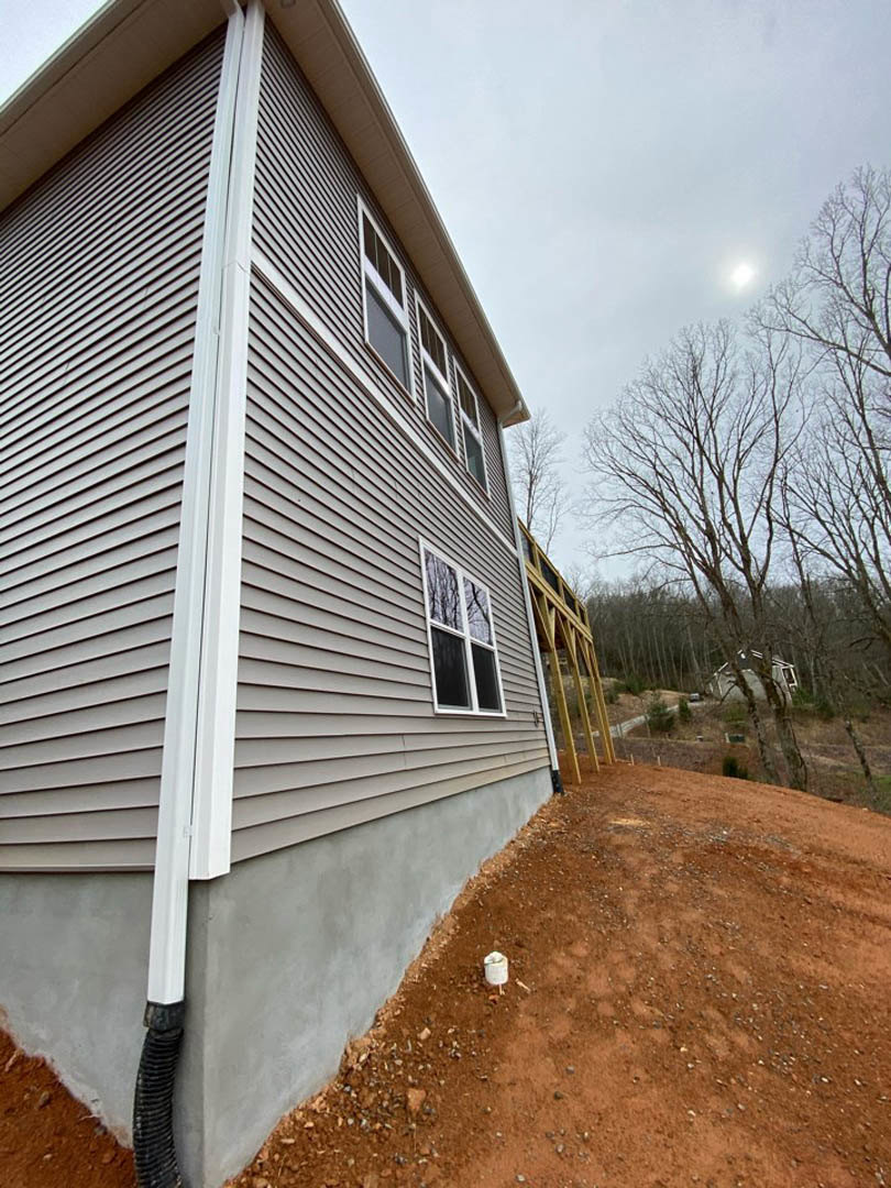 Partially built house with exposed wooden framing, white window trim, unfinished deck, leafless tree nearby, white paint can on bare ground, cloudy sky overhead