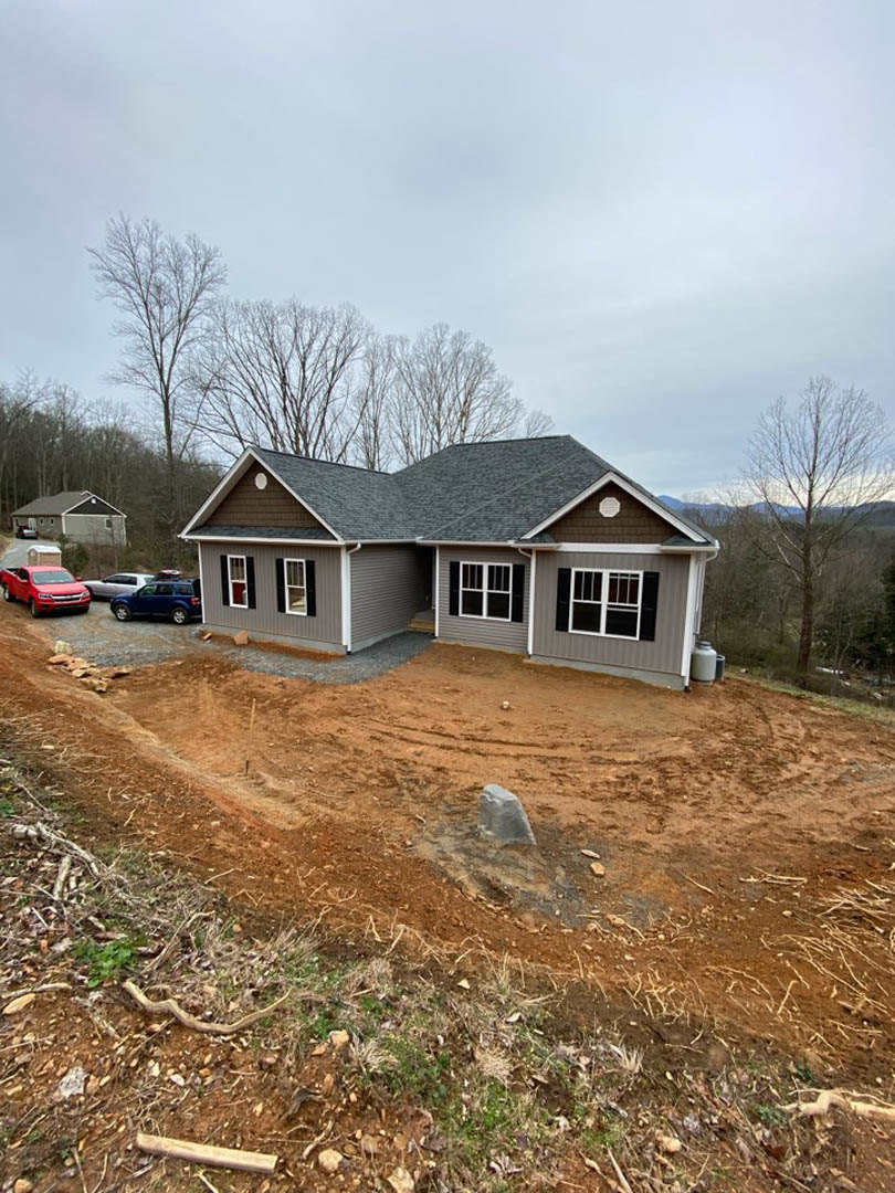 Black roof home with white siding, large front windows, leafless tree nearby, red truck parked on roadside, dirt yard with scattered rocks, cloudy sky overhead.