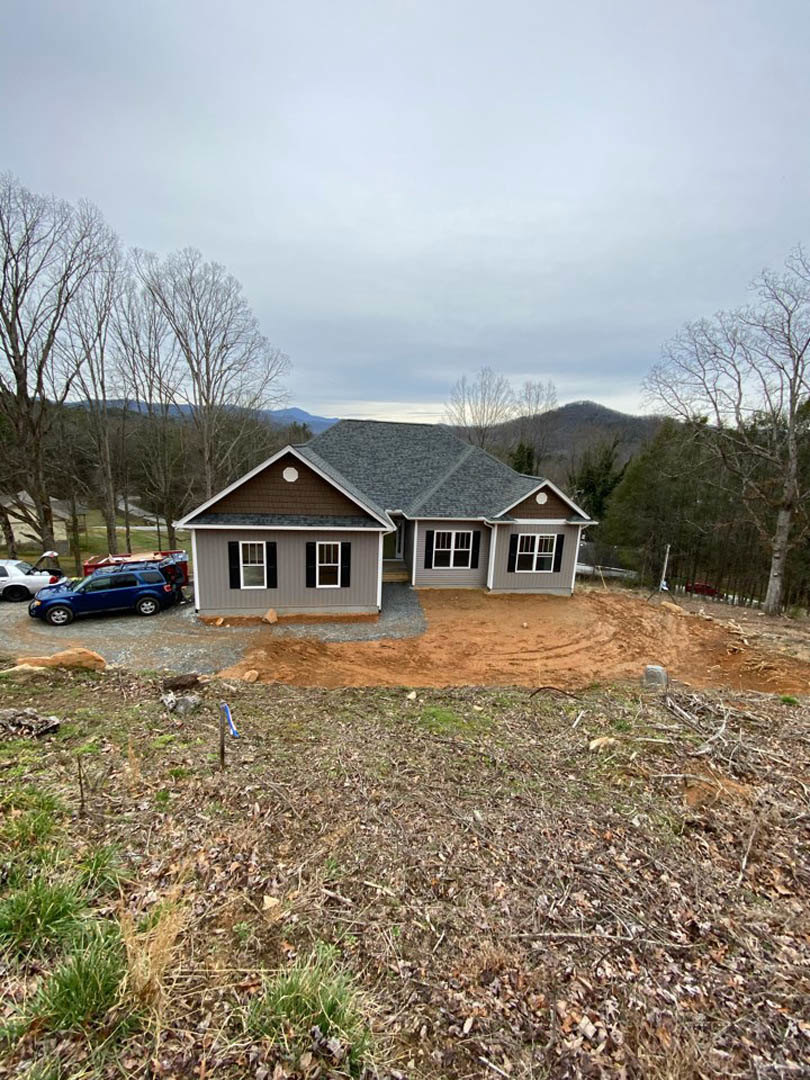 Two-story house with grey roof, light siding, large driveway, blue car parked in front, trees and grass surrounding property, cloudy sky overhead