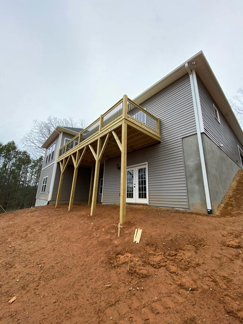 Two-story house with light-colored siding, wooden deck, covered front porch, and surrounding trees; pile of soil near foundation.