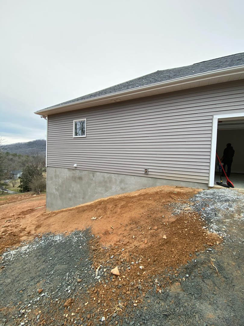 Grey siding home with white garage door, man standing inside garage, pile of dirt and gravel on driveway, window reflecting nearby tree.