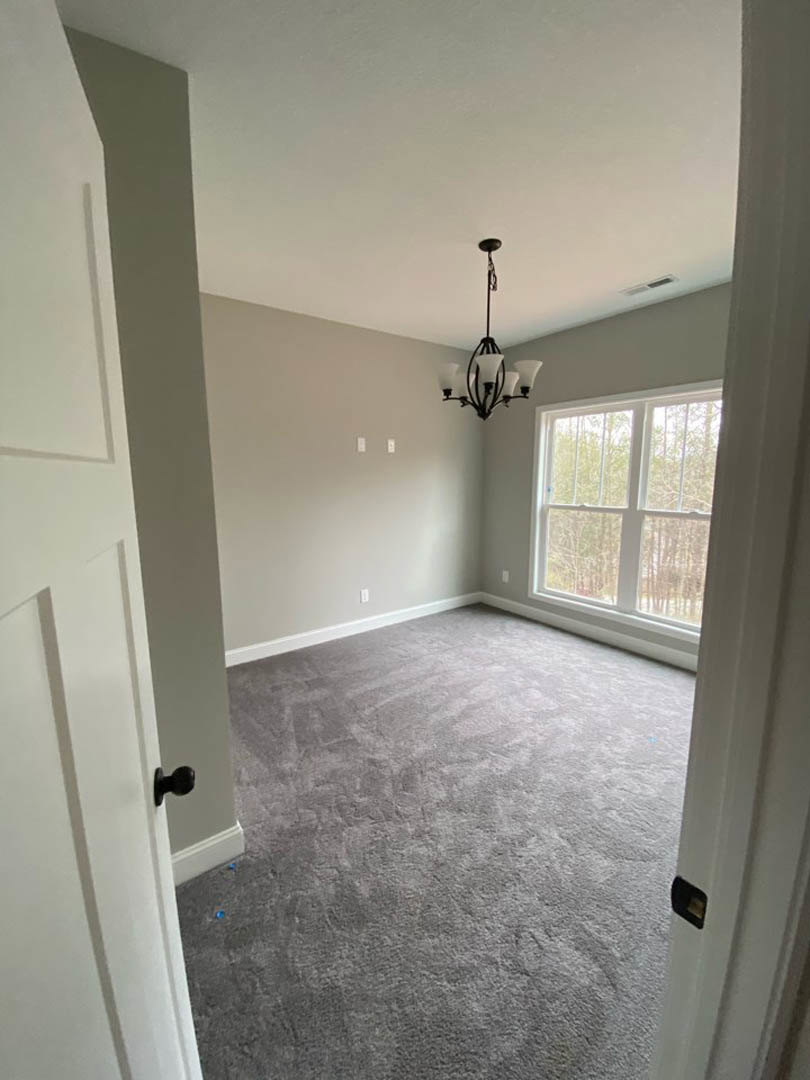 Chandelier hanging from white ceiling above carpeted floor, large window with white frame overlooking trees, neutral walls and grey surfaces throughout room