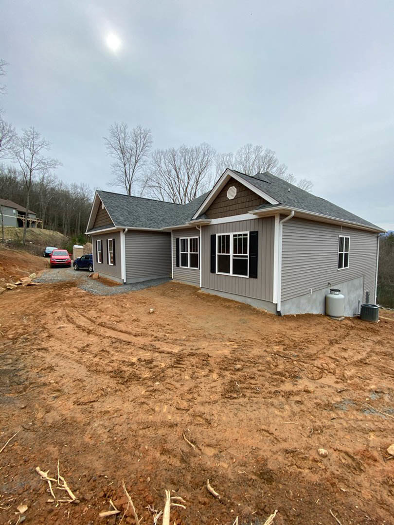 Grey-sided house under construction with black shuttered windows, white container with black lid near dirt patch, car parked in front, cloudy sky and trees in background