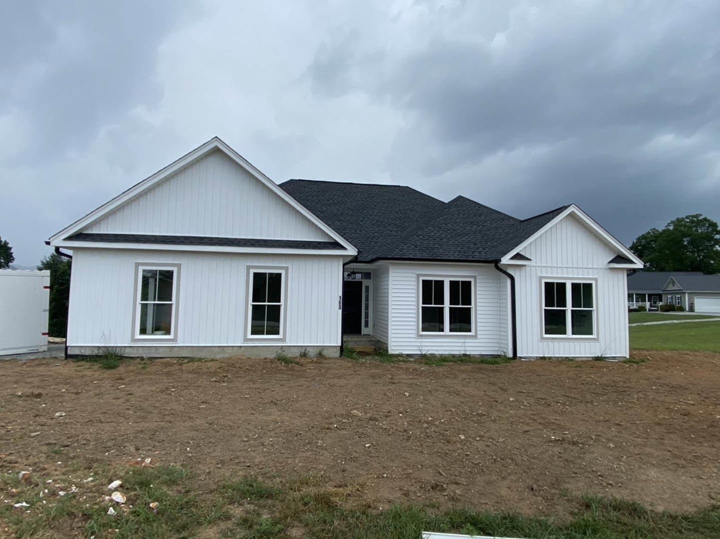 White house with black roof, white-framed windows, and dirt yard with sparse grass under cloudy sky