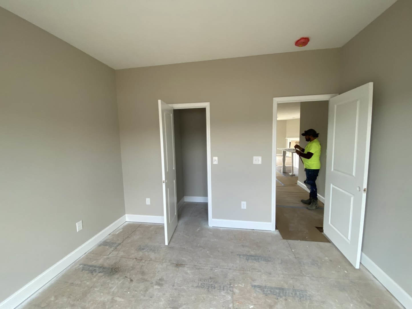 Man in yellow shirt standing on light wood floor between two white doors, white plaster walls, ceiling fixture, red oval object with yellow dots on floor.