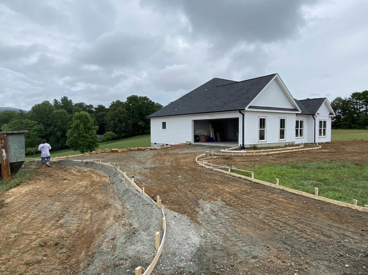 Partially built house with exposed framing, attached garage, dirt driveway, surrounding trees, and cloudy sky