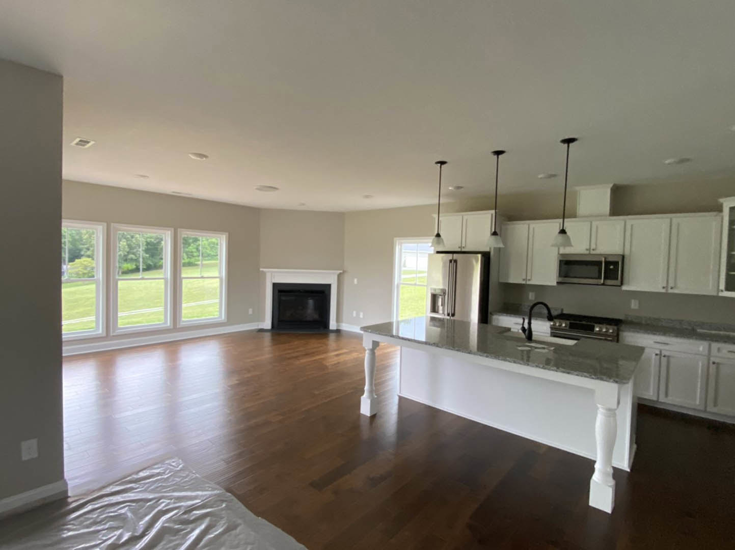 Open-concept kitchen and living room featuring hardwood floors, white cabinetry, kitchen island with built-in sink, stainless steel refrigerator, microwave oven, and a row of large