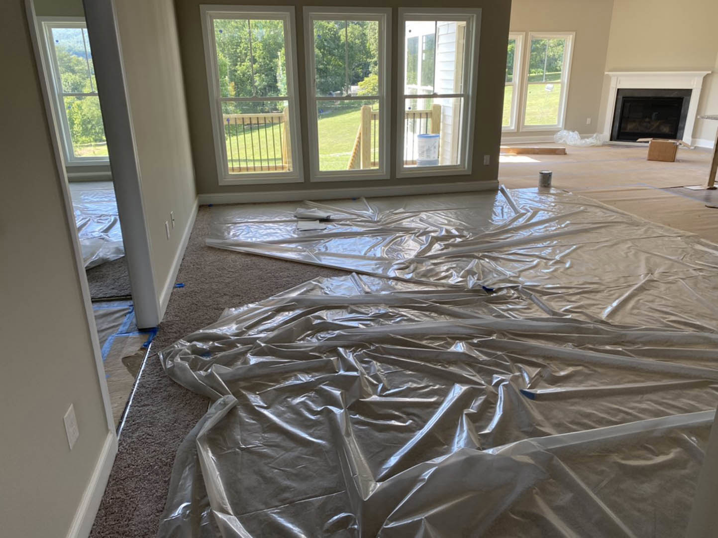 Living room under renovation with plastic sheeting covering the floor, a fireplace with unfinished plaster, large window overlooking deck, brown box on table, and construction