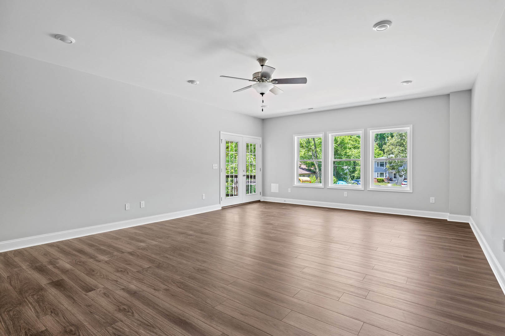 Hardwood floor room with ceiling fan, white double glass doors, row of windows showing trees, white walls and plaster ceiling
