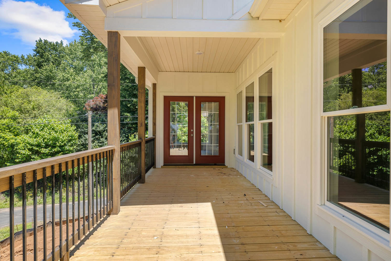 Red double doors with glass panes on a wooden porch, white railing, window overlooking trees, blue sky with clouds above.