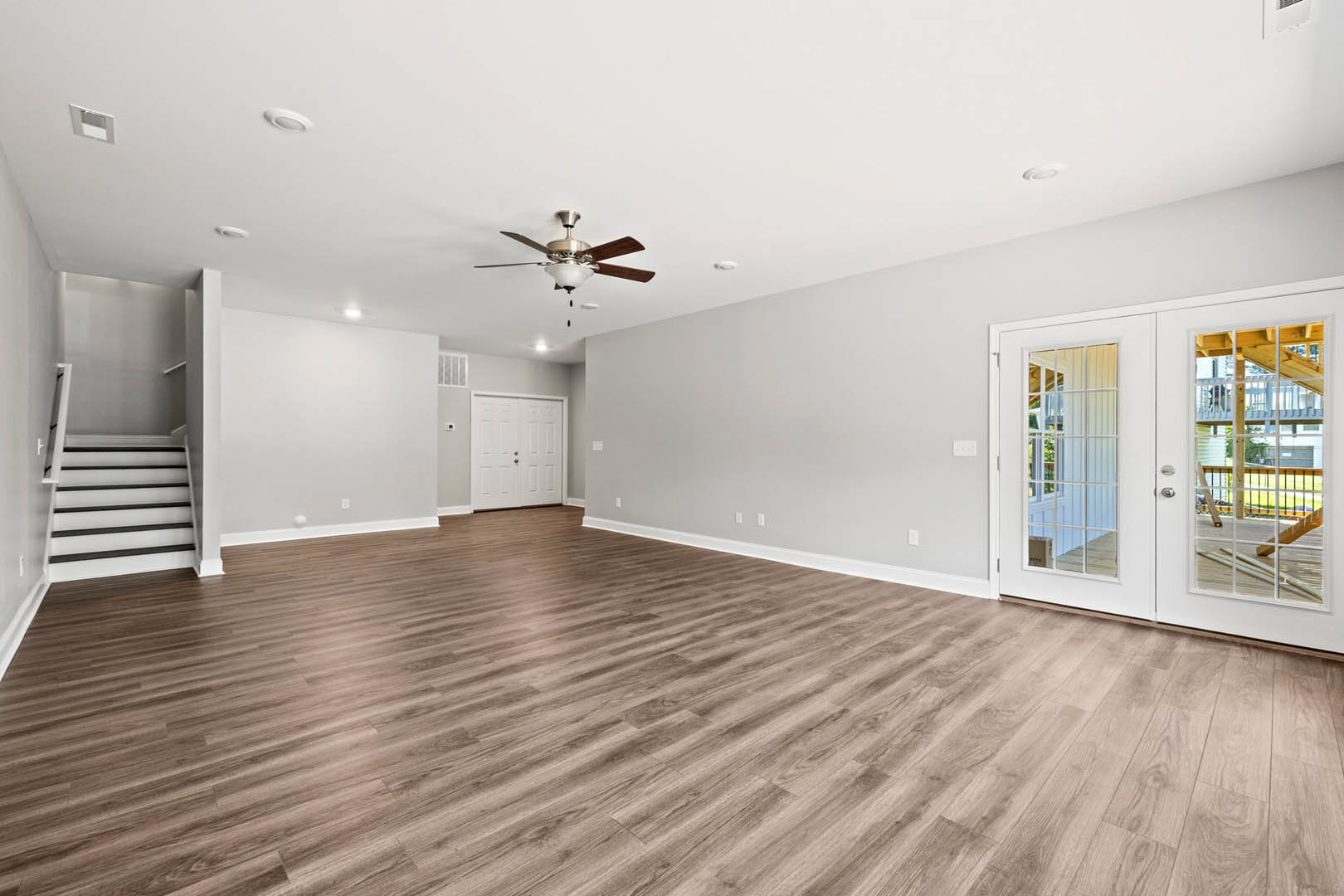 Wood flooring in a bright room with a ceiling fan and light, white double doors with glass panes, plaster walls, and a window overlooking a neighboring house