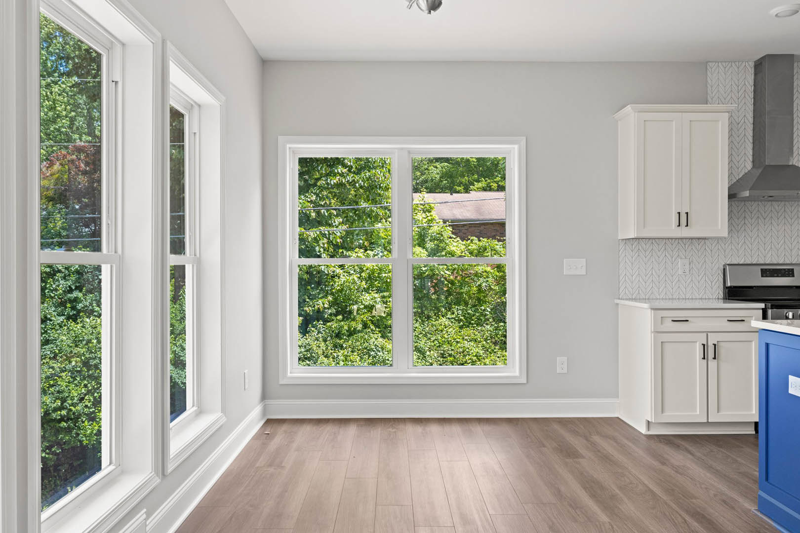 Hardwood floor room featuring a large window with tree views, white walls, white cabinetry with black handles, and a blue door