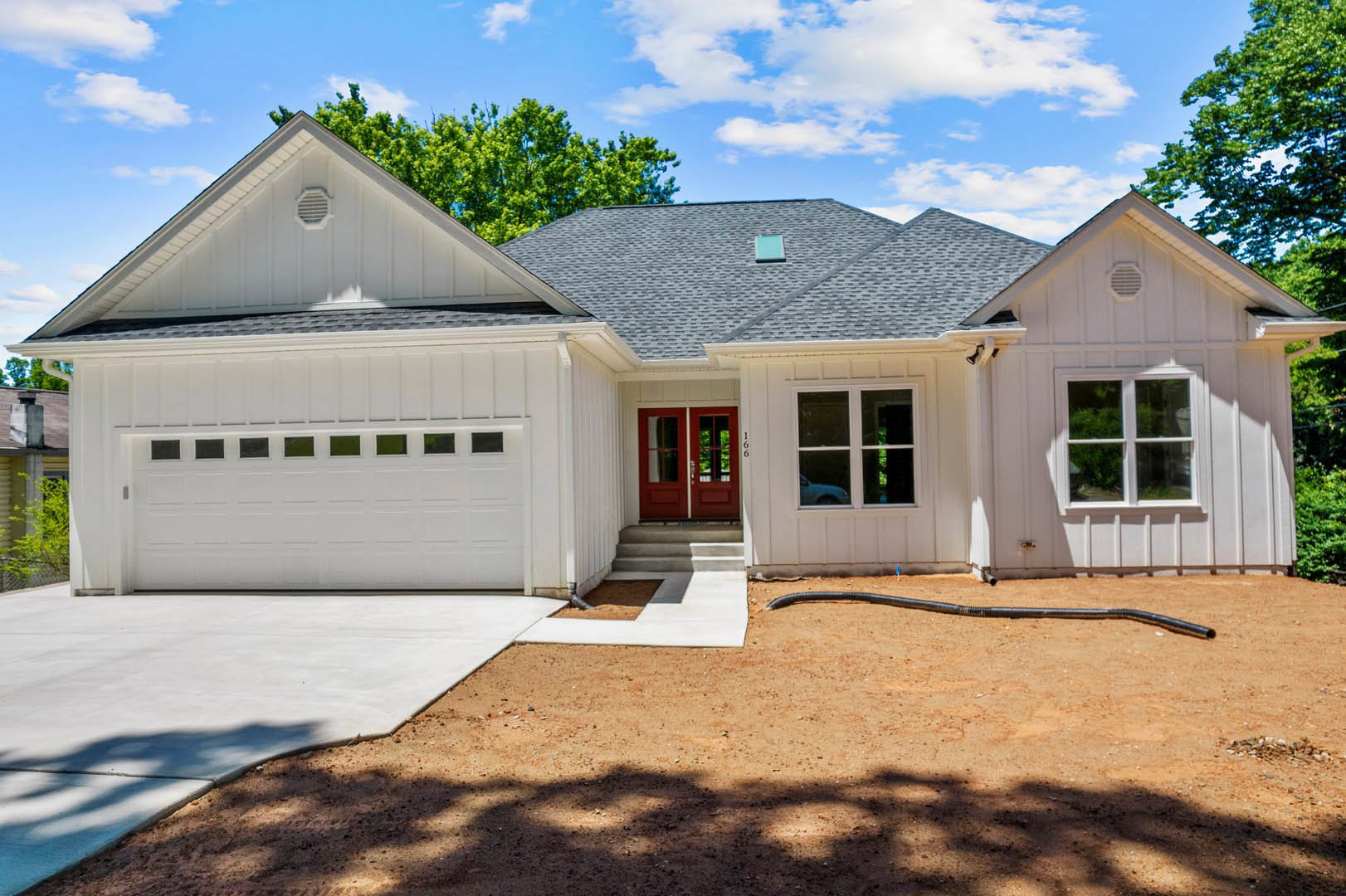 Two-story house with gray siding, white trim, attached garage, concrete driveway, and prominent red front door; large windows and small porch visible, surrounded by landscaped yard