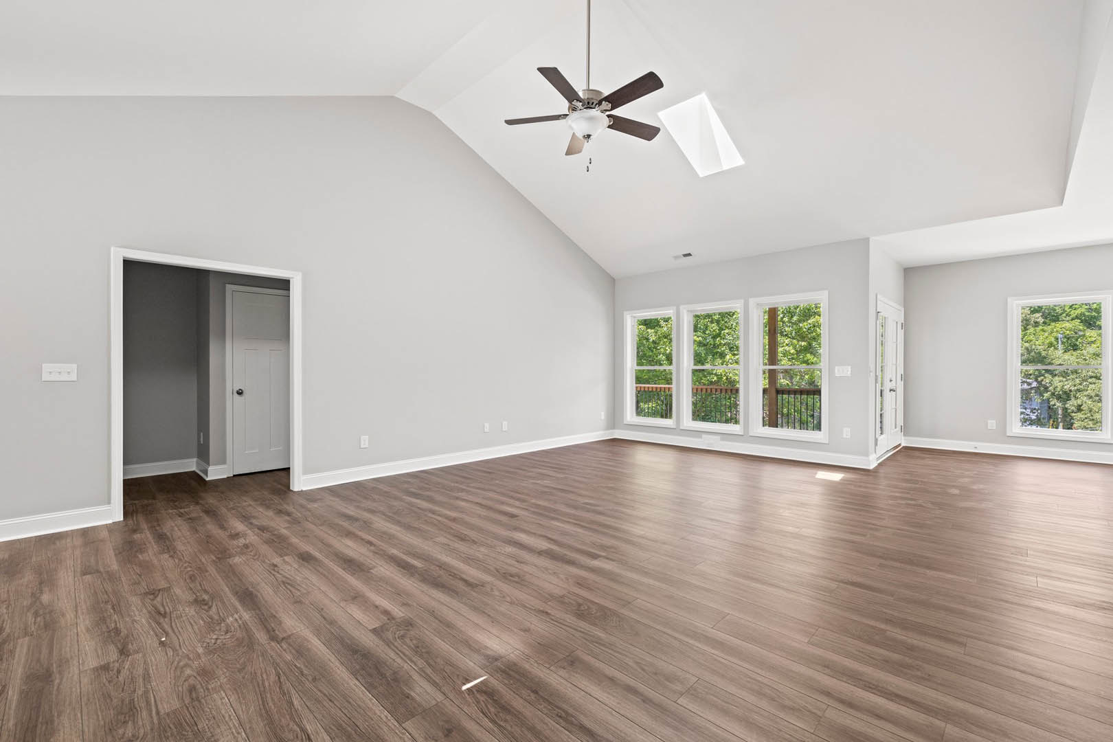 Spacious room featuring hardwood floors, white walls, ceiling fan with light fixture, large window overlooking trees, and white door with black knob