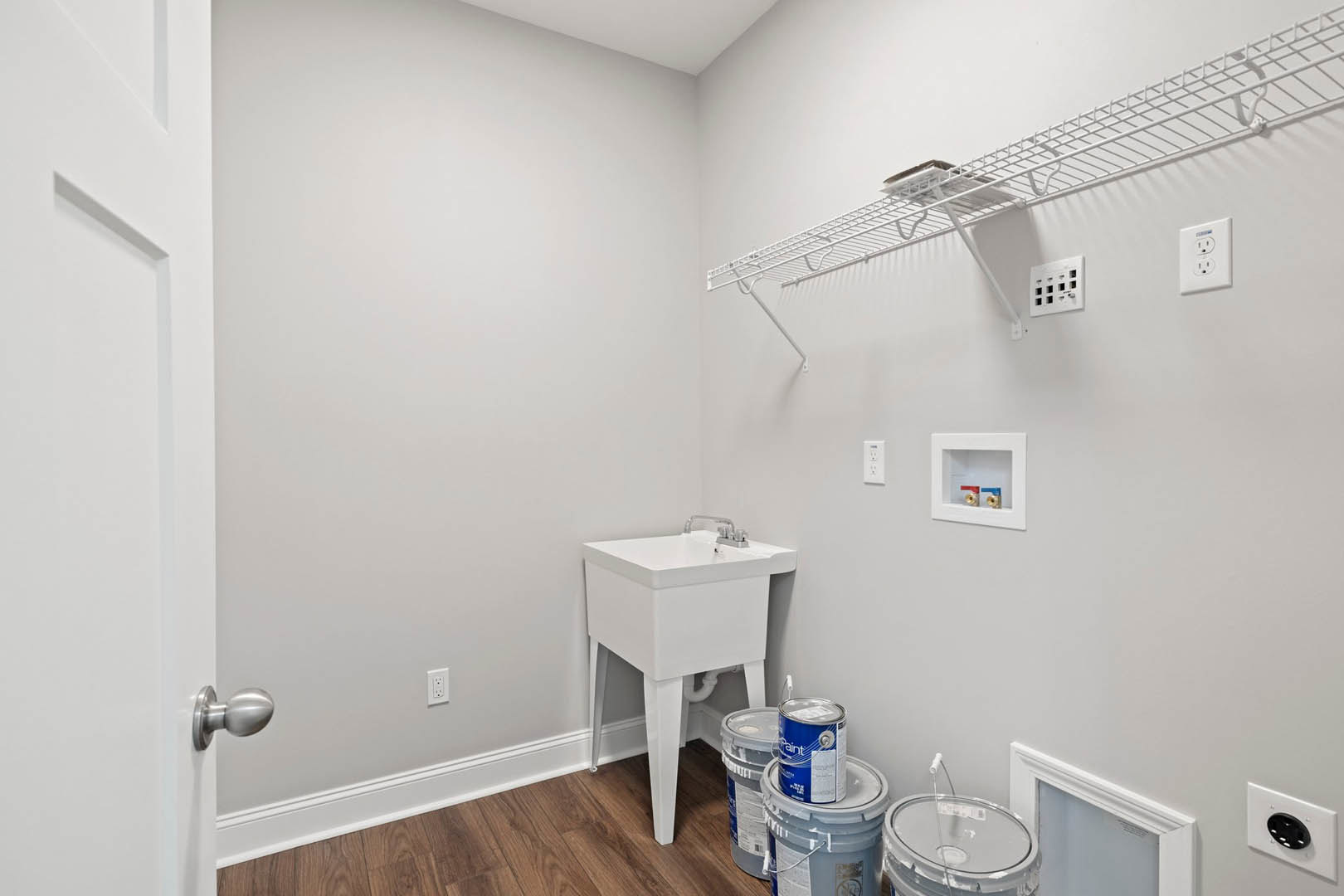 White-painted utility room with a wall-mounted sink, paint buckets on the floor, white shelving, electrical outlet, and a window providing natural light.