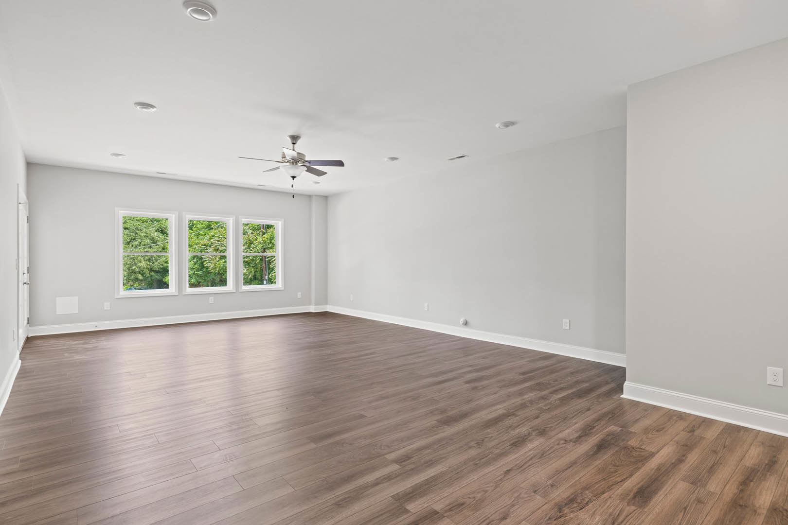 Hardwood floor room with white walls, black trim, ceiling fan with light, row of windows overlooking trees