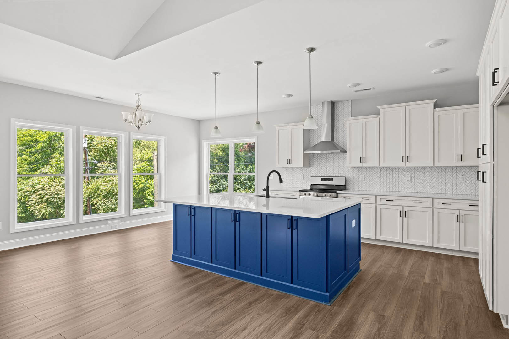 Blue kitchen island with white countertop, matching blue cabinetry, wood flooring, large window showing trees outside, modern chandelier overhead, stainless steel sink and
