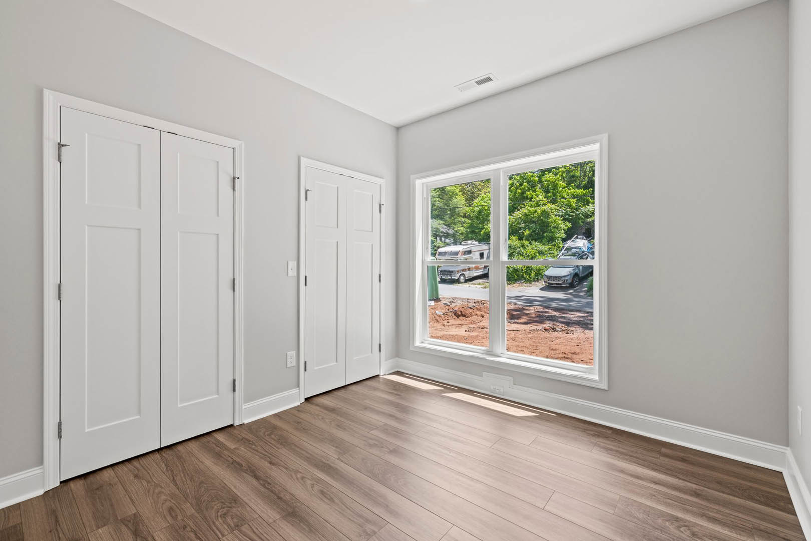 Room with white closet doors featuring black handles, wood flooring, white cabinets, and a window overlooking a road with cars