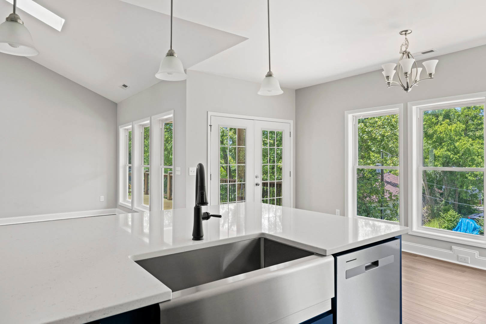 Modern kitchen featuring white cabinetry, stainless steel sink and dishwasher, glass-paneled double doors, large window with tree views, recessed lighting in a white ceiling, and