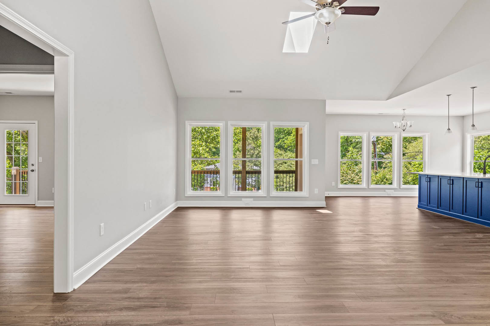 Spacious room with wood flooring, ceiling fan, multiple windows overlooking trees, blue cabinets with white countertops, glass-paneled white door, and interior railing.