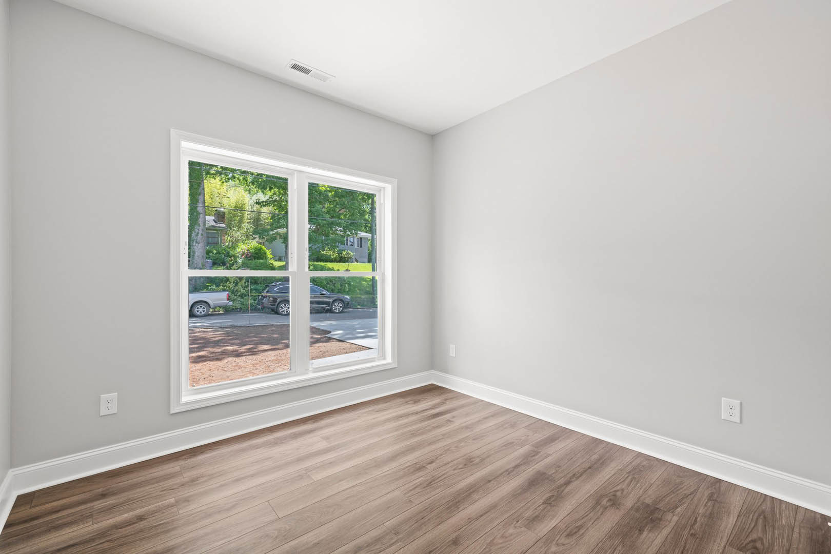 Sunlit room with large window overlooking street and parked cars, hardwood floor with white baseboard trim, white electrical outlet on plaster wall