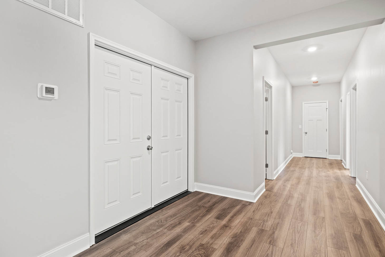Hallway with white paneled doors featuring silver knobs, wood laminate flooring, white plaster walls, and a digital thermostat mounted near the entry.