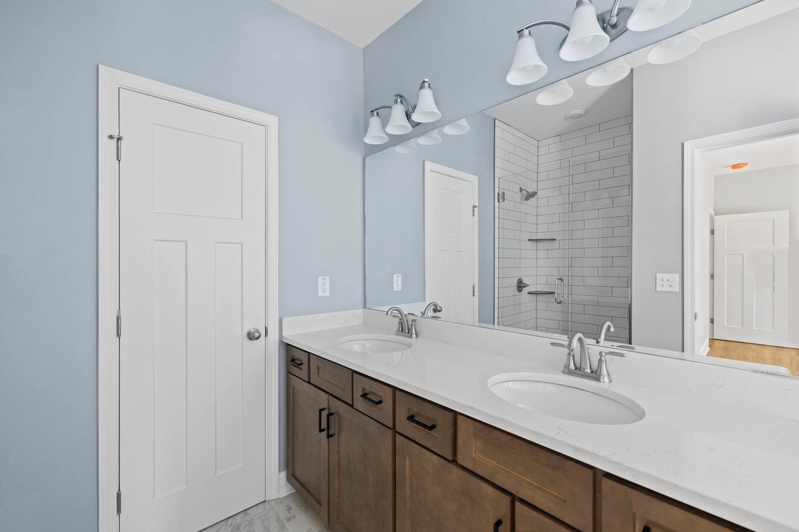 Bathroom with double vanity featuring undermount sinks, quartz countertop, large frameless mirror, glass-enclosed shower with tile walls, white cabinetry, silver hardware, and