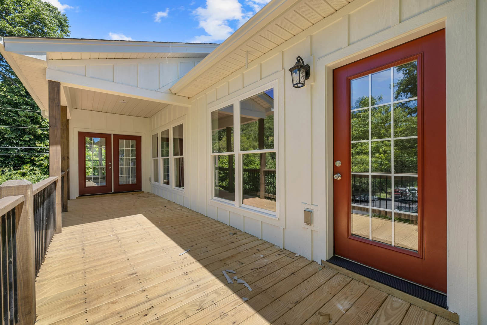 White siding house with red framed double doors and windows, wood deck porch, outdoor lamp, tree shade, and blue sky in background