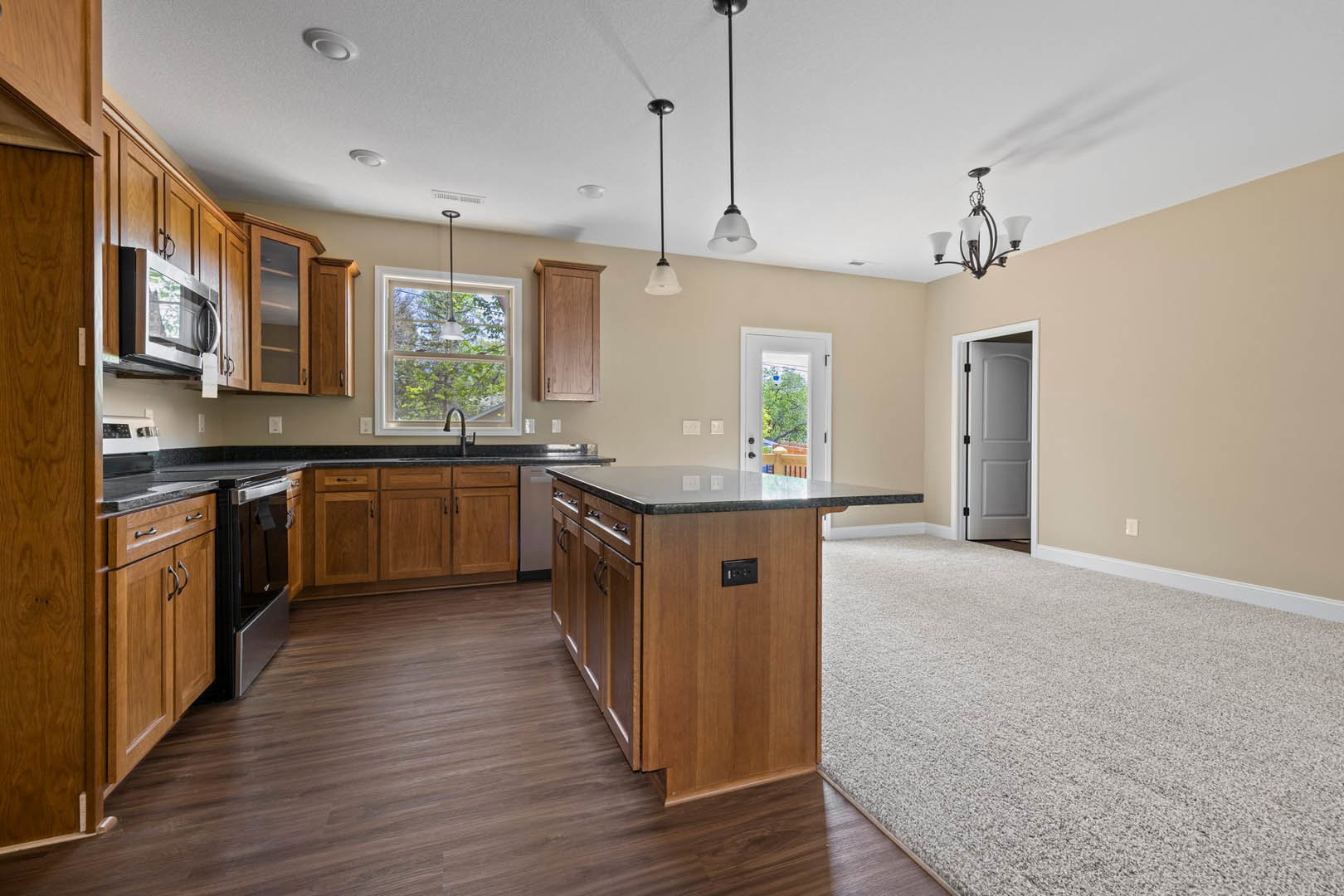 Kitchen with wood cabinets, black stove, open microwave, black countertop island, ceiling light, white door with black handle, carpeted adjacent room