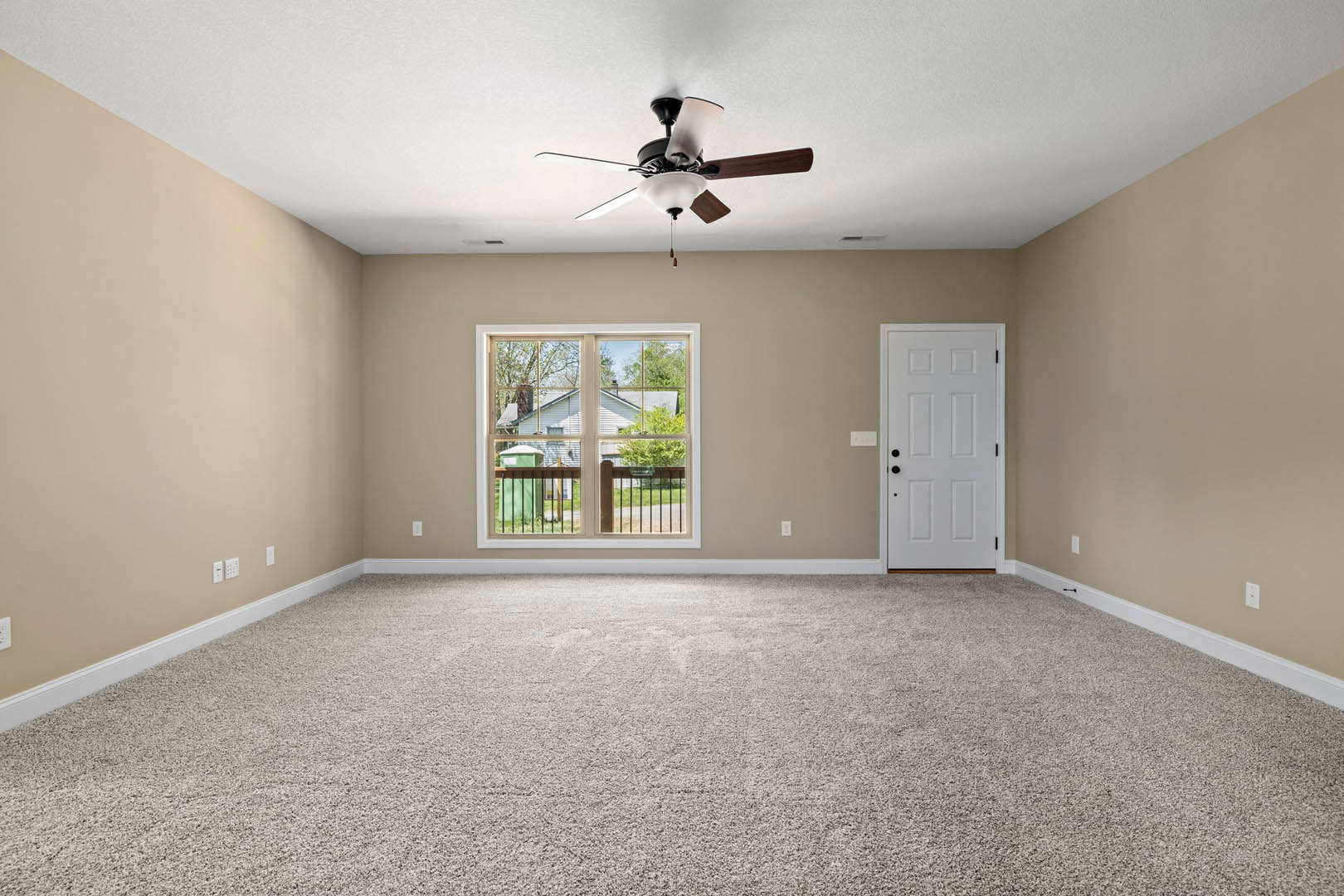 Bedroom with beige carpet, white walls, ceiling fan with light fixture, large window overlooking house and trees, white door with black hardware, smoke detector on ceiling.