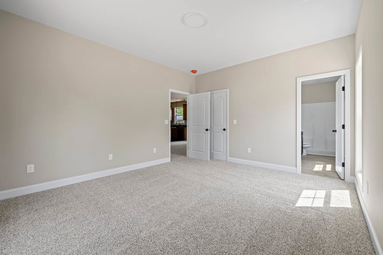 Carpeted bedroom with white walls, open white door featuring black knobs, adjacent bathroom with white wall, white square tiles on grey countertop