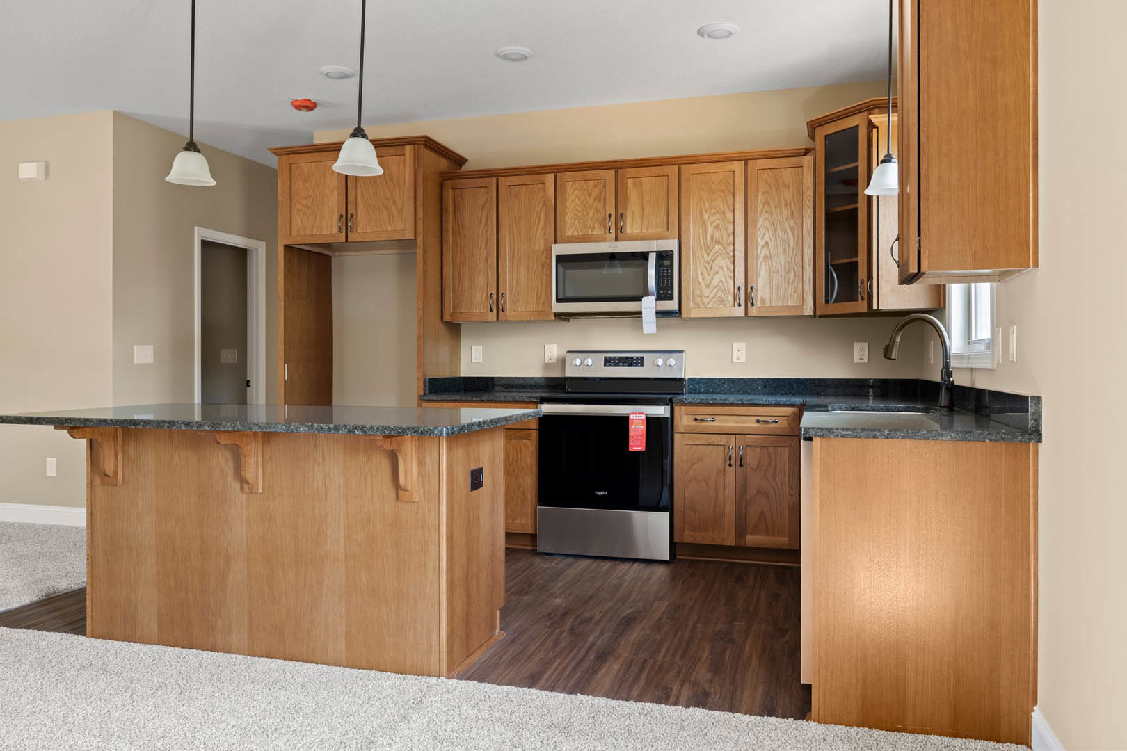 Kitchen featuring wooden cabinets, black countertop, stainless steel oven with red tag, glass-door microwave, white-framed brown wall, and close-up of wooden door and metal pole