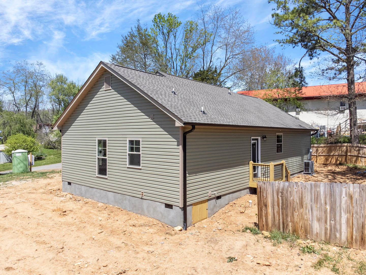 Two-story house with white-framed windows, wooden deck and porch, fenced backyard, green container with white lid, grey rectangular planter, surrounded by trees and plants under a