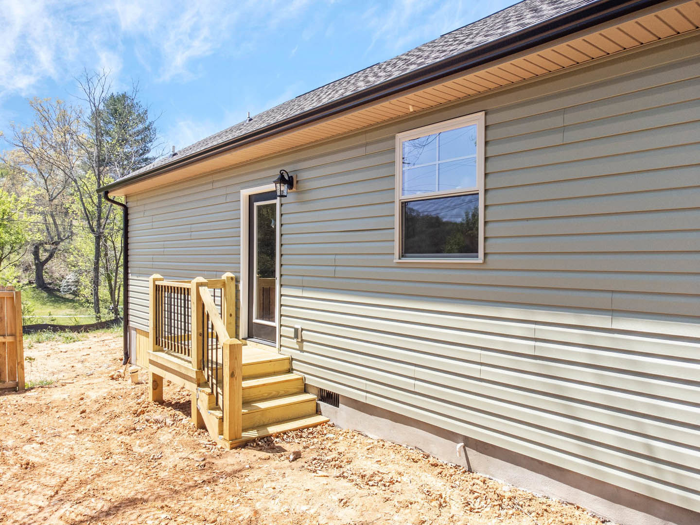Two-story home with light siding, wooden porch and staircase, metal railing, and trees in the background