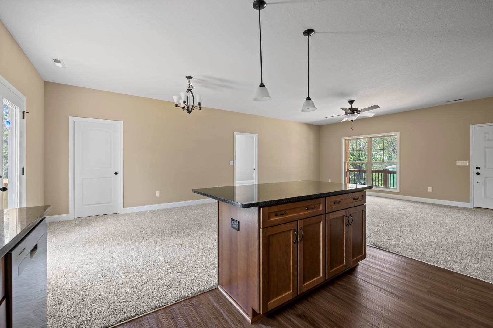Kitchen with black countertops, white cabinetry, ceiling fan, kitchen island, white door with black handle, window overlooking trees, light wood flooring