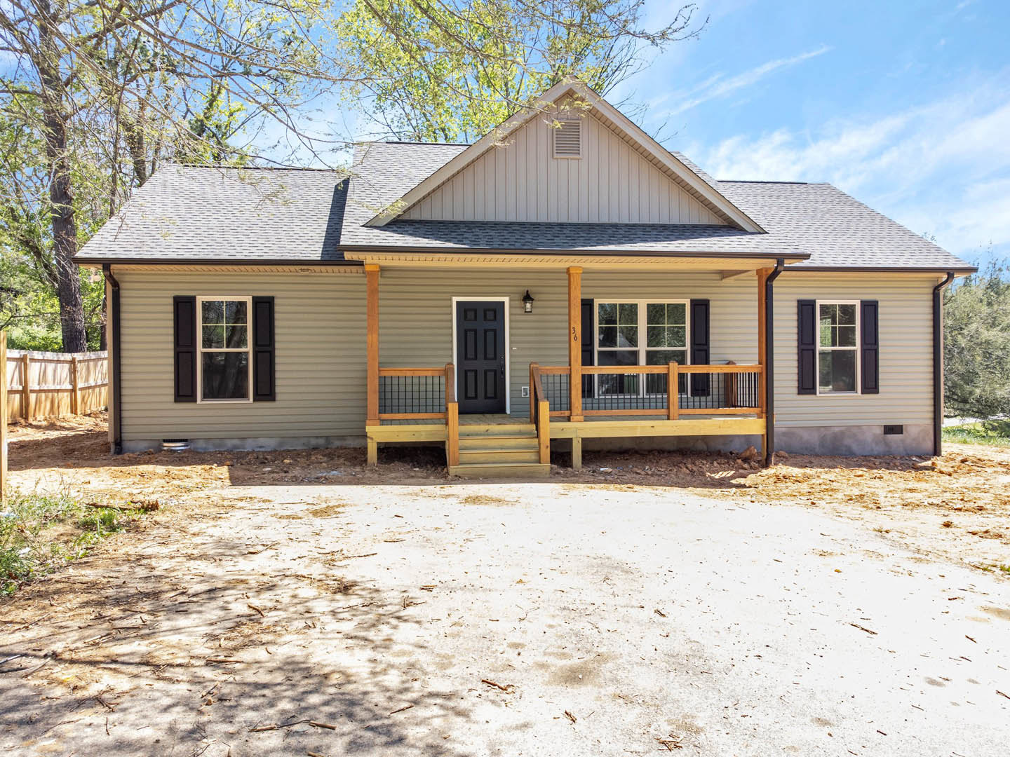 Two-story house with white siding, black front door, wooden porch featuring a bench, white-framed windows, dirt driveway, and surrounding trees.
