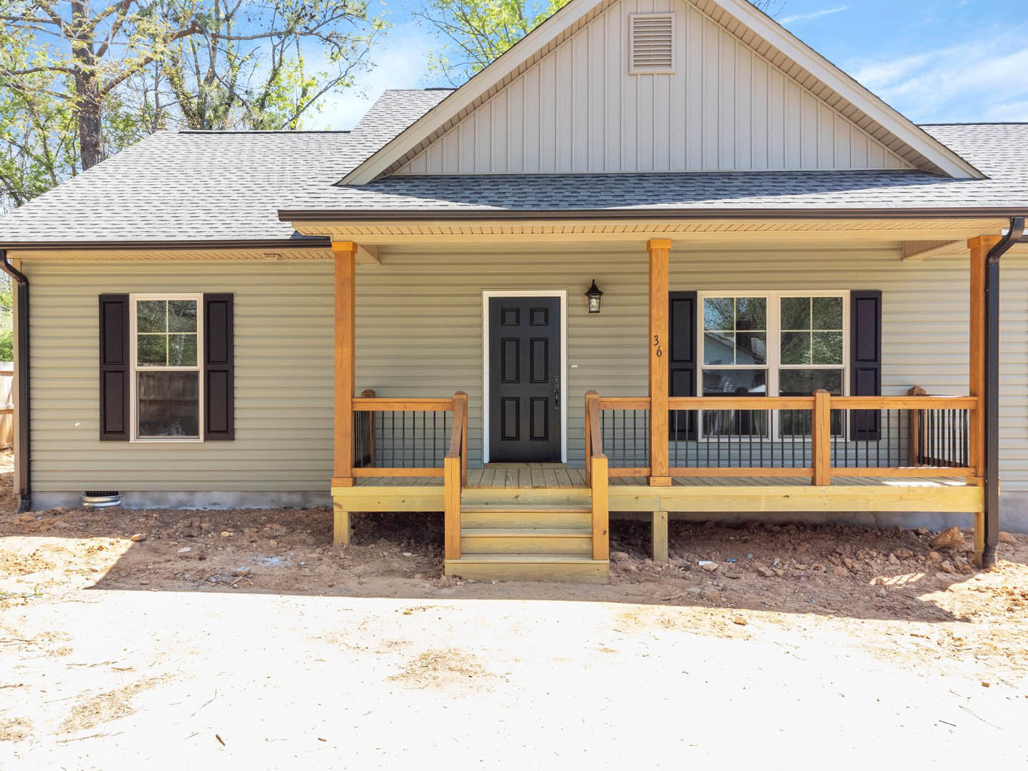Two-story home with gray siding, black front door, white-framed windows, covered porch, wooden deck with metal railing, and surrounding trees