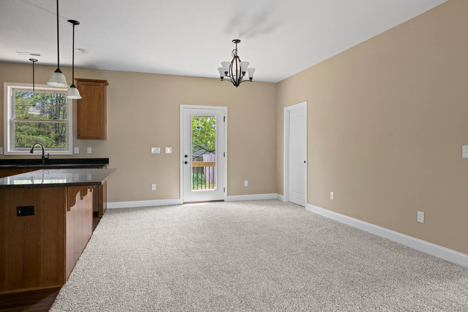 Open kitchen with white cabinetry, stone countertops, stainless steel sink beneath a window, adjacent carpeted room with white door featuring black knobs, modern chandelier