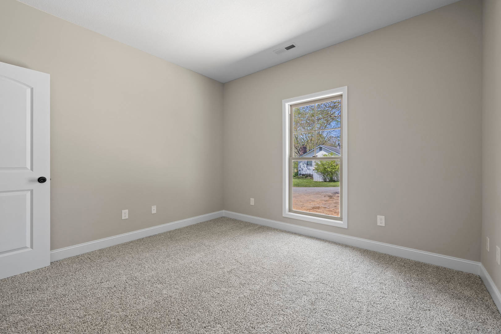Carpeted room with white walls, large window overlooking neighboring house and trees, close-up of white door and wall molding