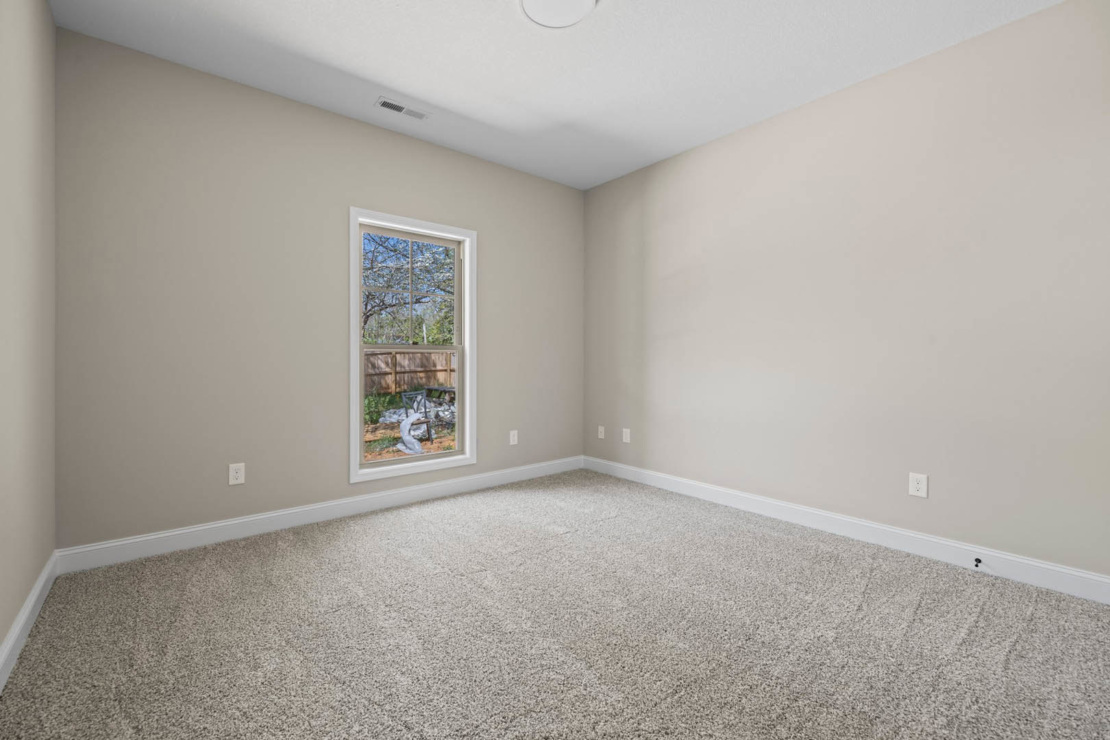 Carpeted room with white walls, large window overlooking backyard with trees, white ceiling featuring round flush light, single chair with plastic bag placed near window